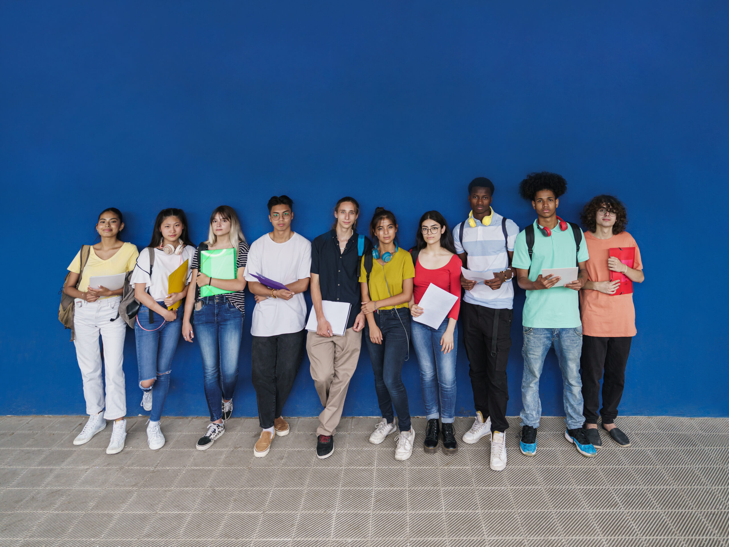 Group of multiracial teenager students looking to camera standing over blue wall copy space. Teenage multi ethnic classmates friends diversity in education